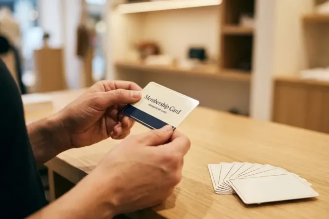 Handen houden een glanzende lidmaatschapskaart vast boven een retailtoonbank, met een waaier van identieke kaarten ernaast in crème-, marineblauw- en goudtinten.