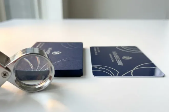 Twee lidmaatschapskaarten naast elkaar: één met matte premium afwerking en reliëftextuur, de ander dunner met vlakke print, met een loupe erbij.