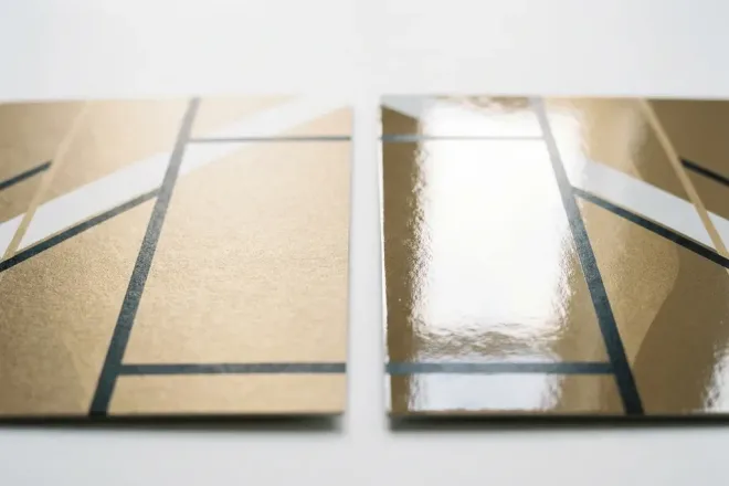 Twee visitekaartjes naast elkaar op wit studiooppervlak, één met matte en één met glanzende afwerking, verlicht om textuurcontrast te benadrukken.