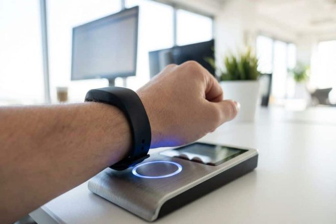 Persoon draagt zwarte RFID-polsband bij moderne kaartlezer met blauwe LED-indicator op wit bureau in kantooromgeving