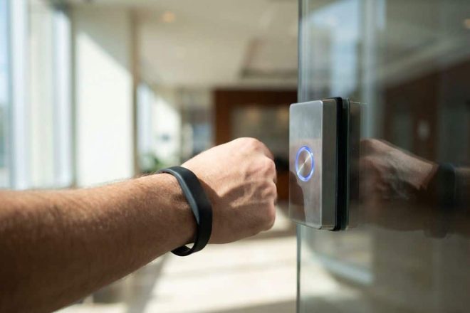 Persoon met zwarte RFID-polsband die toegangskaartlezer op glazen kantoor deur benadert, LED-indicator gloeit blauw