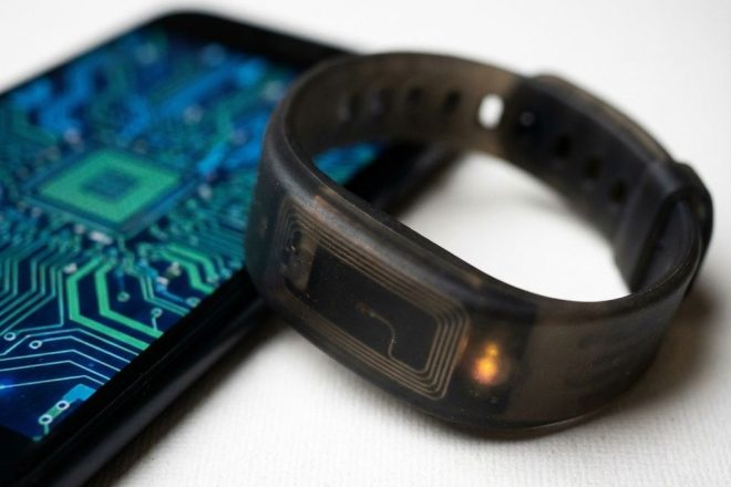 Zwarte RFID-polsband met amber LED-indicator naast smartphone op witte ondergrond, antennelijnen zichtbaar door siliconen