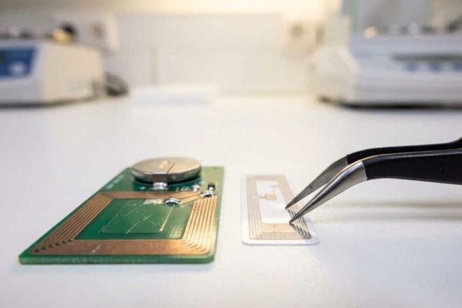 Twee RFID-tags op wit laboratoriumoppervlak, pincet houdt kleinere tag vast, grotere tag toont batterij en antenne