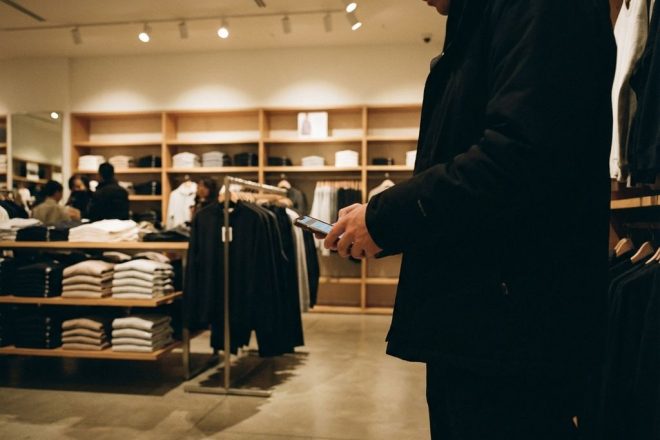 Klant met smartphone bij kledingrek met RFID-tags in moderne minimalistische winkel met warme verlichting