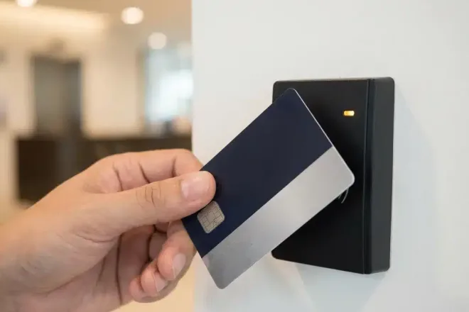 RFID-toegangspas wordt gehouden voor een matte zwarte kaartlezer op een witte muur, met zichtbare chiptextuur en warm omgevingslicht.